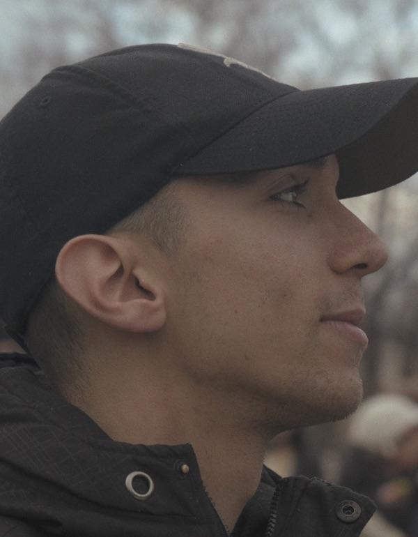 A side-profile close-up of a young man outdoors wearing a black baseball cap and a dark jacket. He looks ahead with a calm, focused expression. The background is softly blurred, suggesting a crowd or public setting behind him.