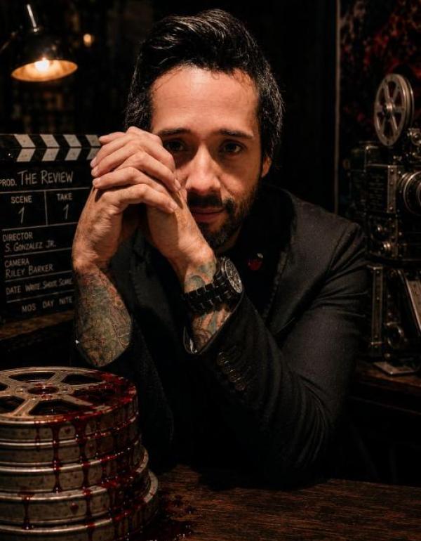 Director Samuel Gonzalez Jr. in a cinematic portrait surrounded by vintage filmmaking equipment and blood-streaked film reels, representing his horror film THE REVIEW.