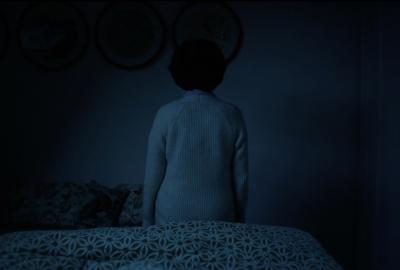 An elderly woman sitting at the edge of a bed, facing away.