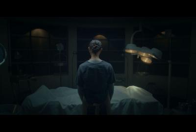 A woman stands in front of a dead body covered with a sheet in a morgue