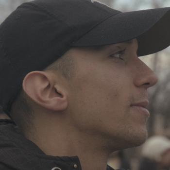 A side-profile close-up of a young man outdoors wearing a black baseball cap and a dark jacket. He looks ahead with a calm, focused expression. The background is softly blurred, suggesting a crowd or public setting behind him.